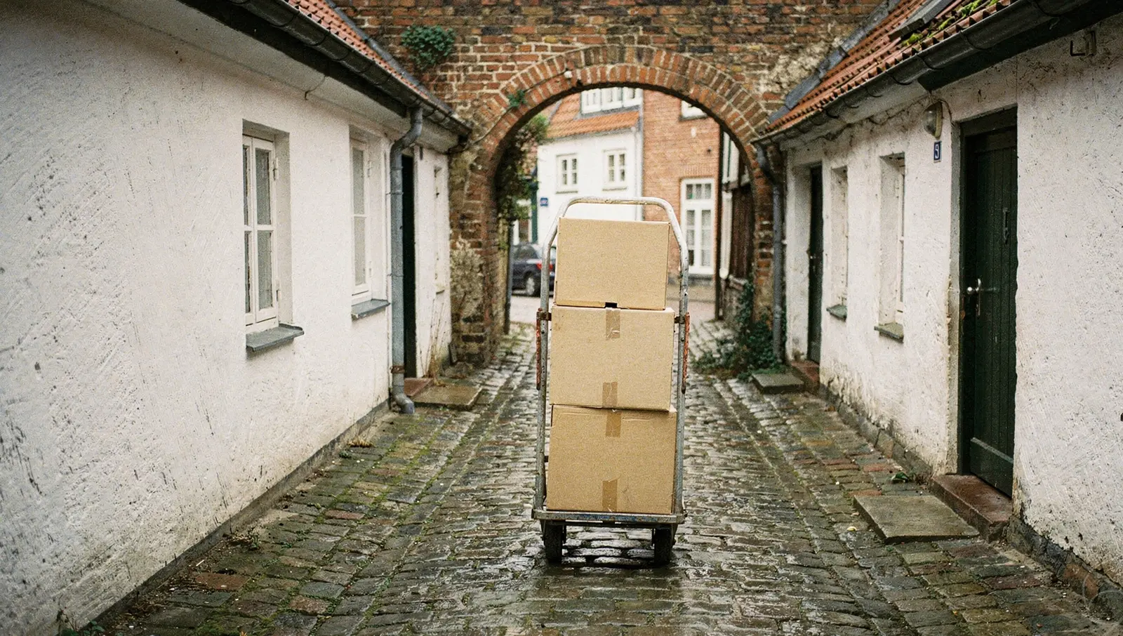 Ein schmaler, kopfsteingepflasterter Lübecker Gang zwischen weiß gekalkten Häuschen, in der Mitte eine Sackkarre mit drei Umzugskartons, hinten ein Backsteinbogen.