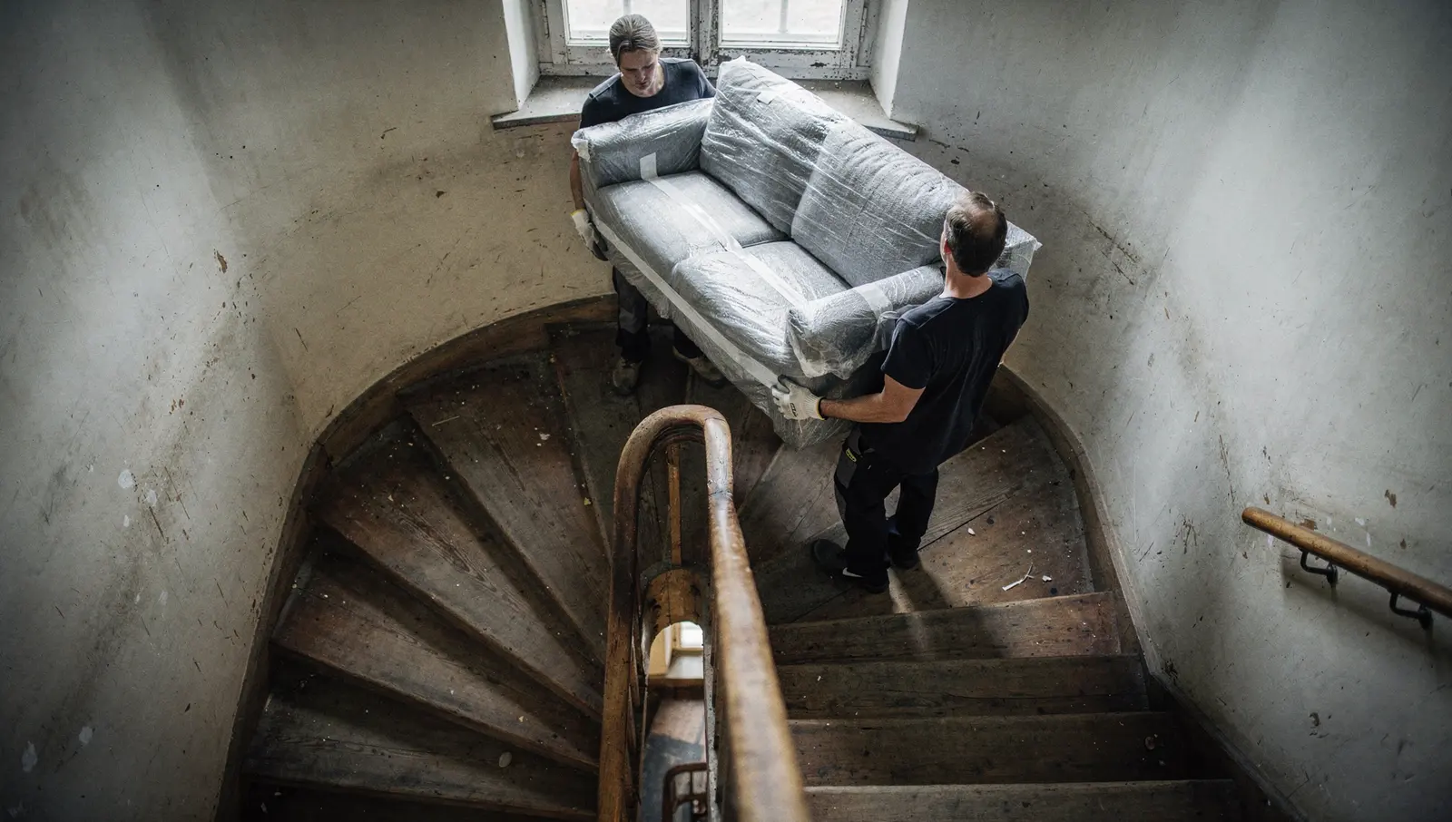 Zwei Möbelpacker tragen ein in Folie gepacktes Sofa eine enge, ausgetretene Holzwendeltreppe hinunter, von oben fotografiert.